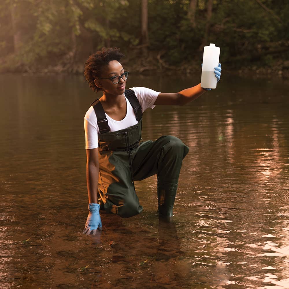 Western student wearing hip waders, taking a water sample from a river.